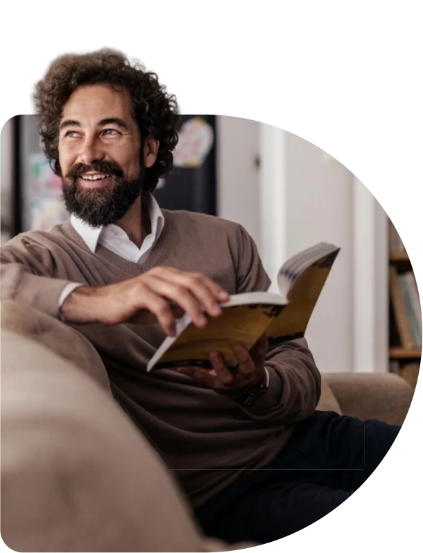Homem sorrindo, sentado no sofá com um livro aberto nas mãos.